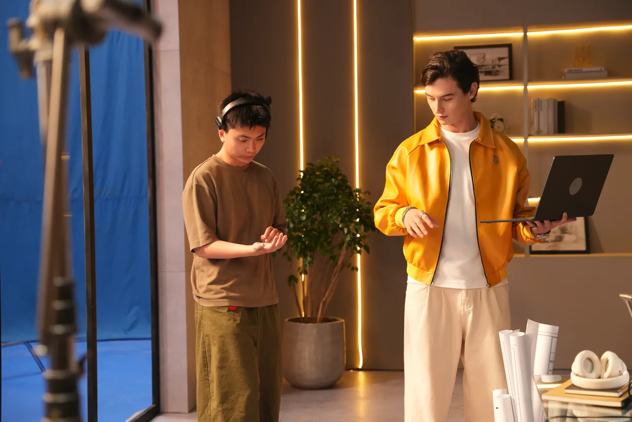 Two performers running a beat beside the plant, the blue screen wrapping in at frame left