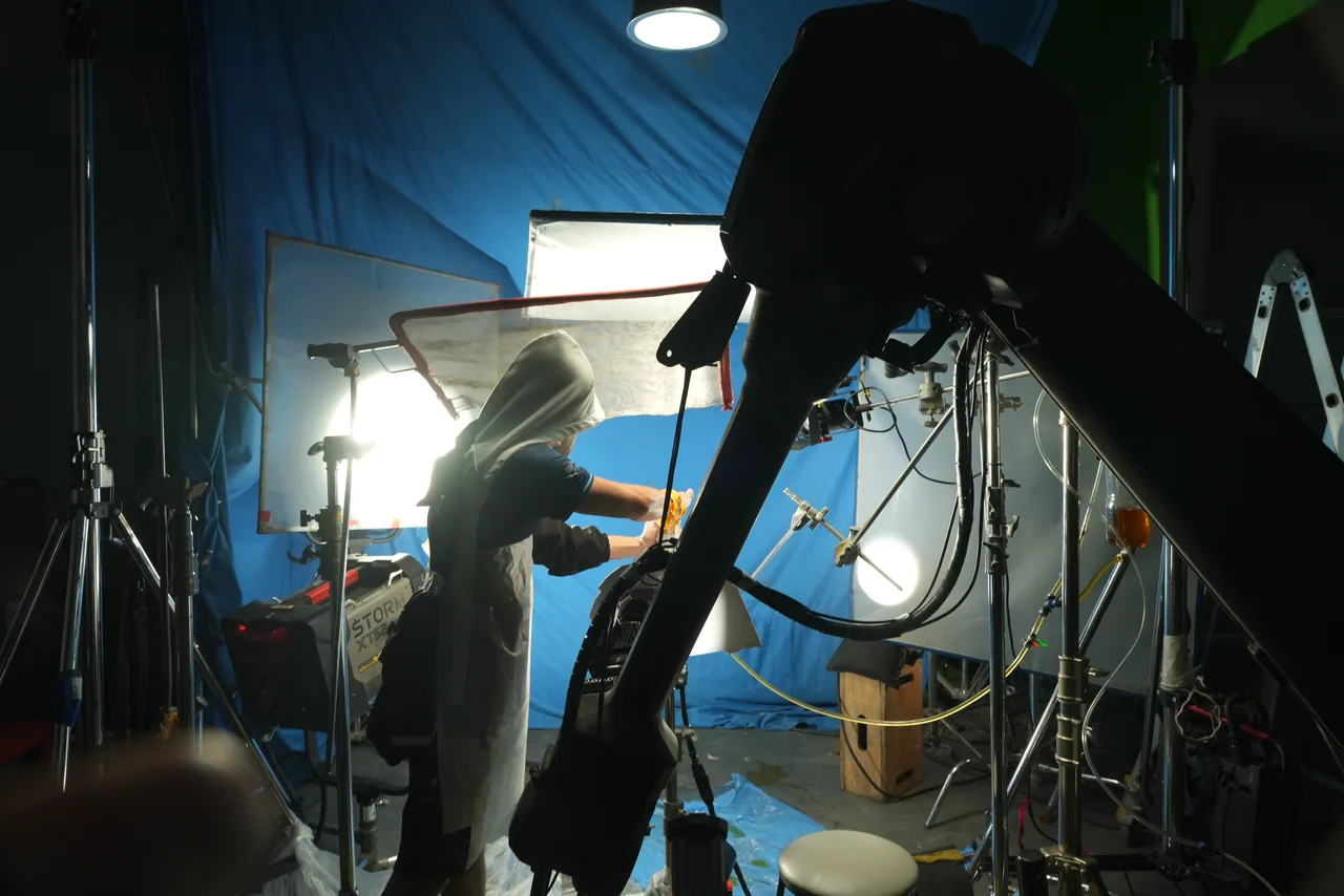 The robotic camera arm silhouetted against a blue-lit stage, a soft box burning through behind it